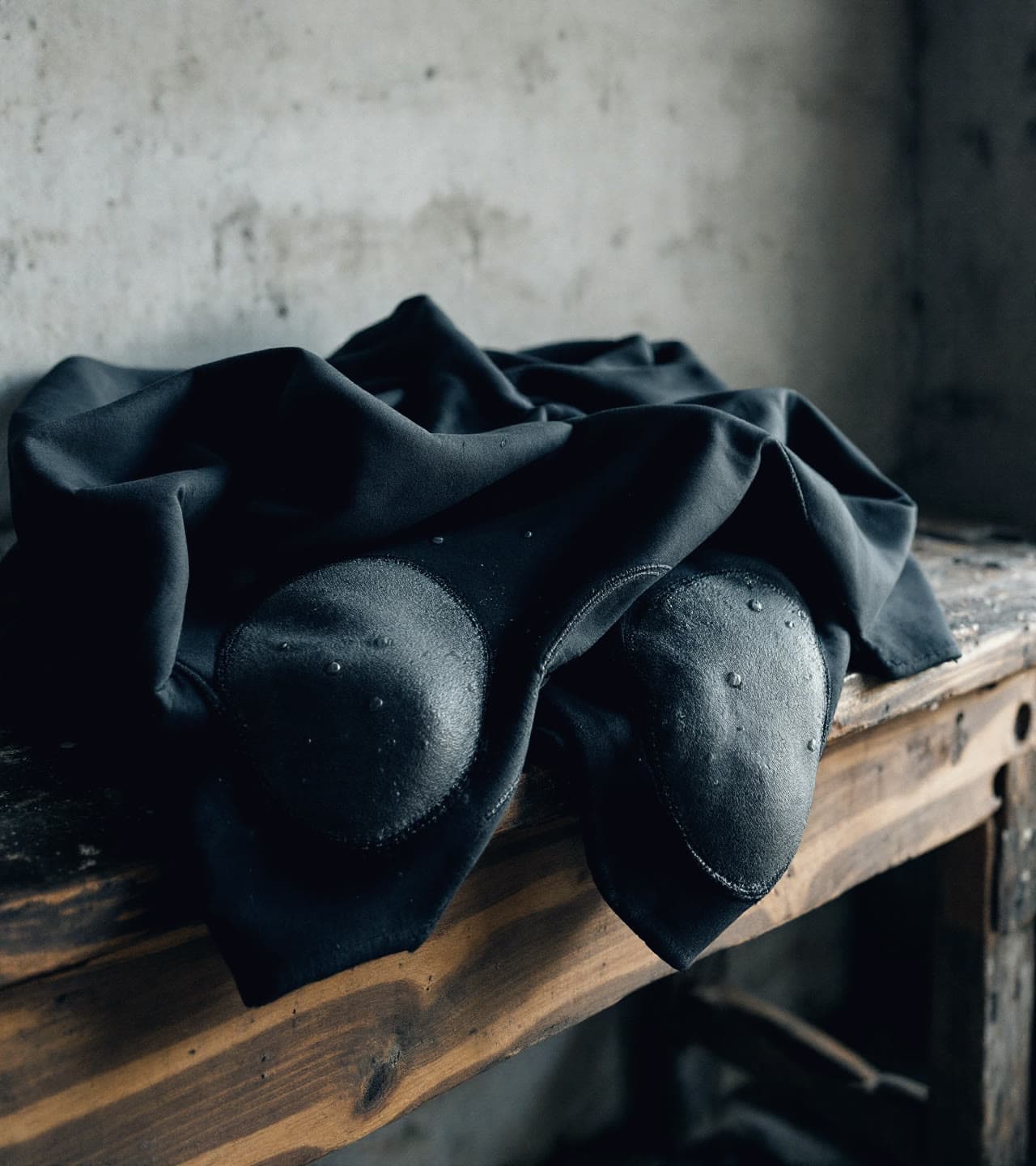 A worn black surf wetsuit folded on a wooden workshop bench, knee panels faded by salt, a few water droplets